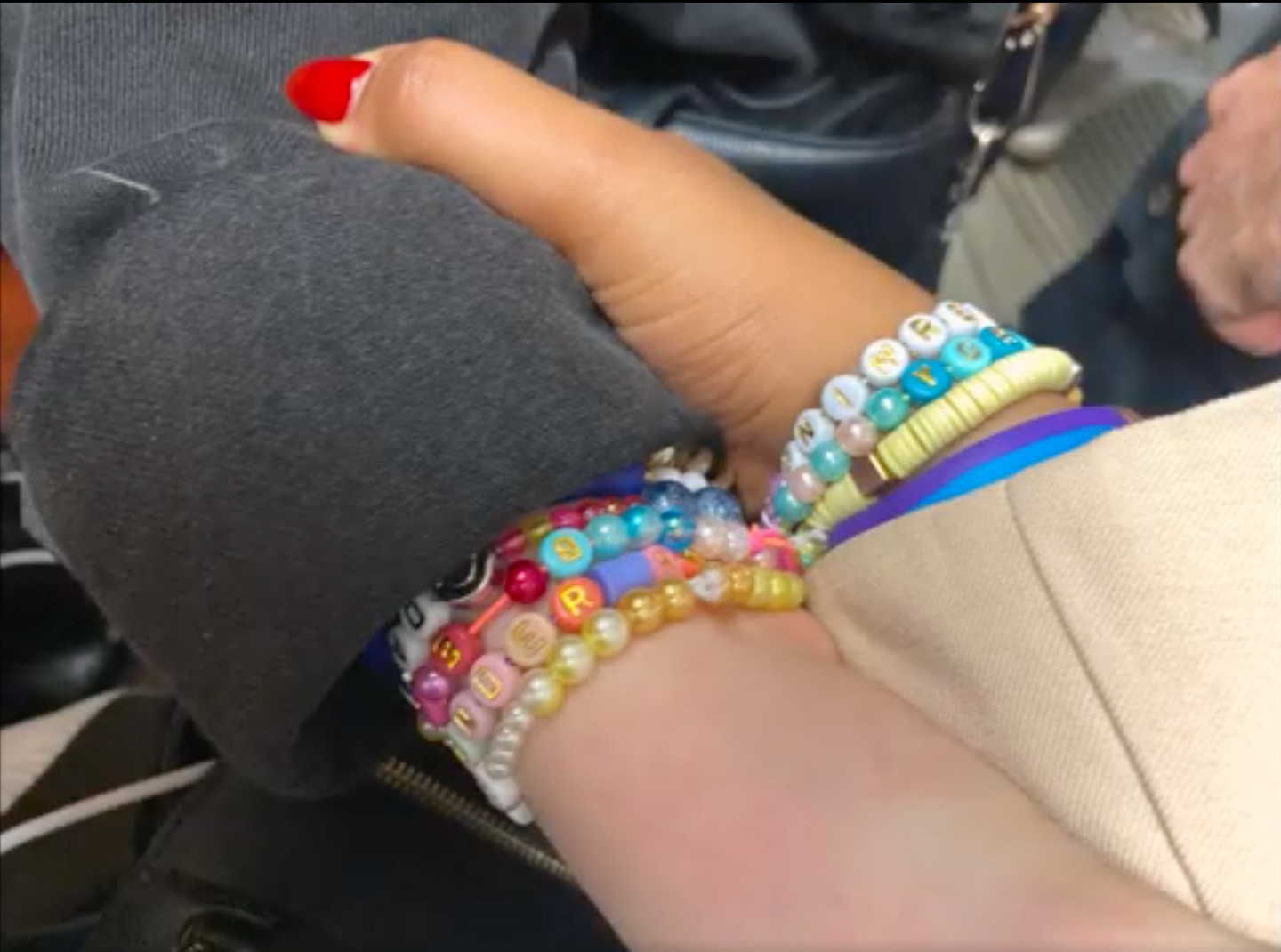 Close-up of a hand with red nail polish wearing multiple colorful beaded friendship bracelets with letter beads on the wrist.