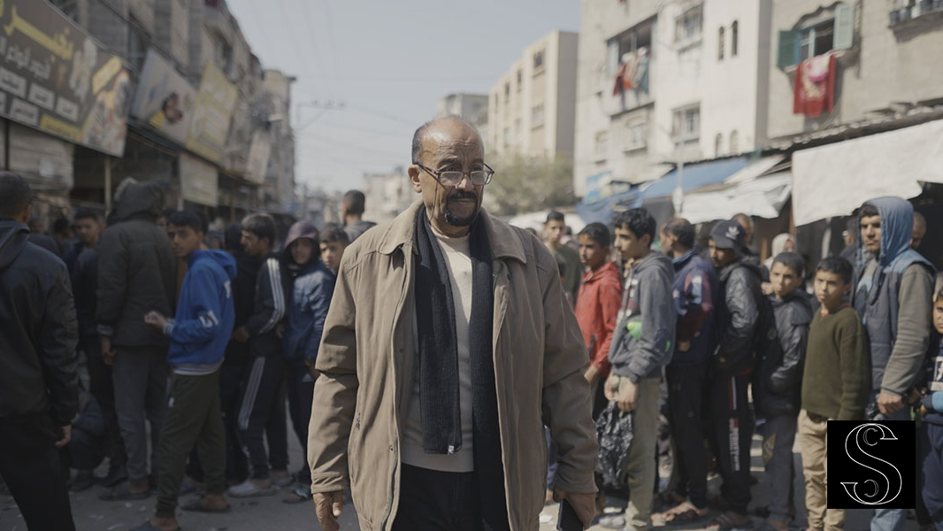 Older man in glasses and tan jacket walks through a crowded urban street lined with buildings, surrounded by onlookers.