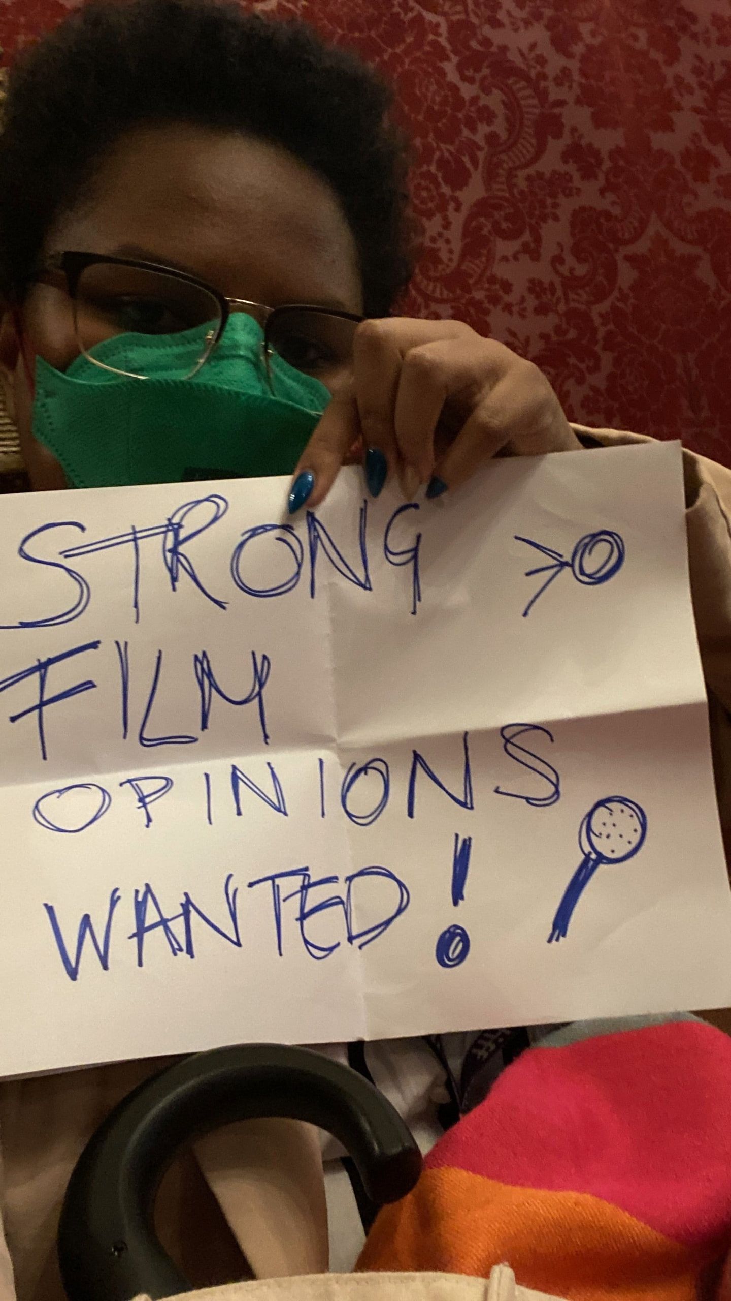 Person wearing glasses and green face mask holds handwritten sign reading "STRONG FILM OPINIONS WANTED!" with microphone doodle.