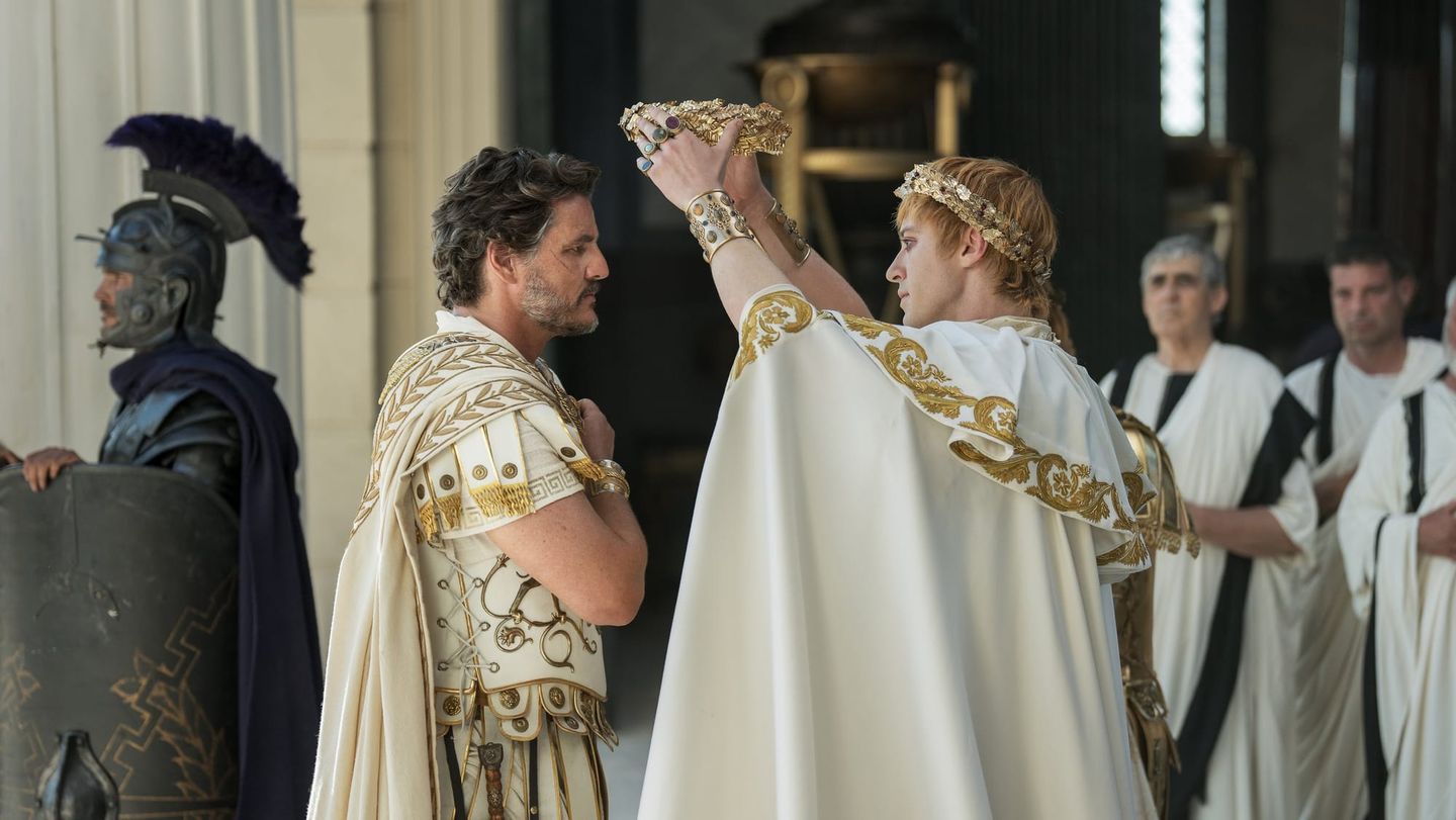 Young man in white Roman emperor robes places a golden crown on a kneeling man in Roman attire, with guards and senators behind.