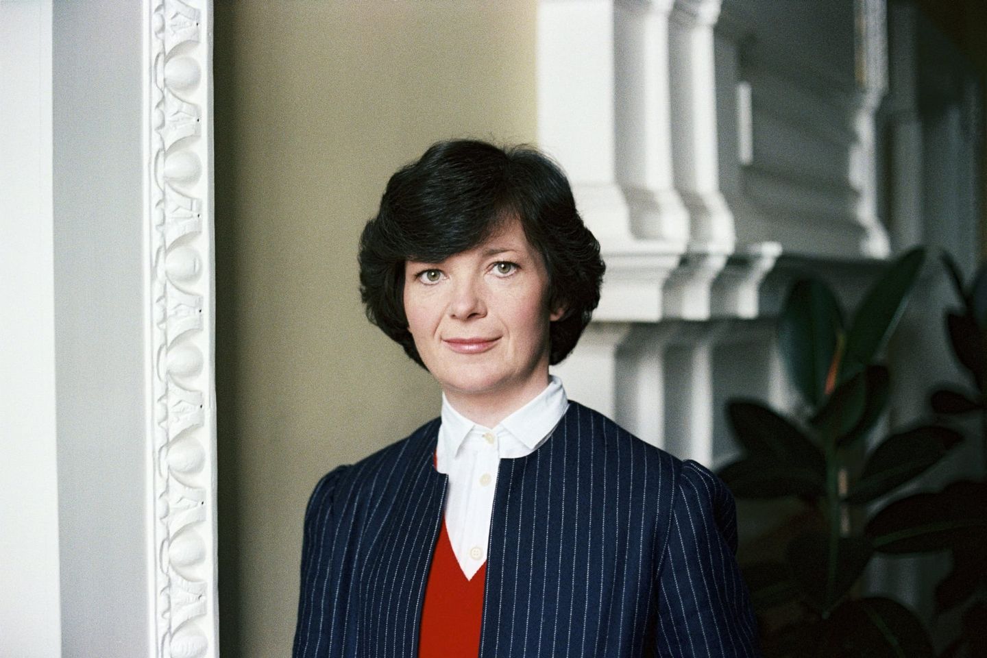 Woman with short dark hair wearing a navy pinstripe blazer, red sweater, and white collared shirt, standing by ornate white molding.