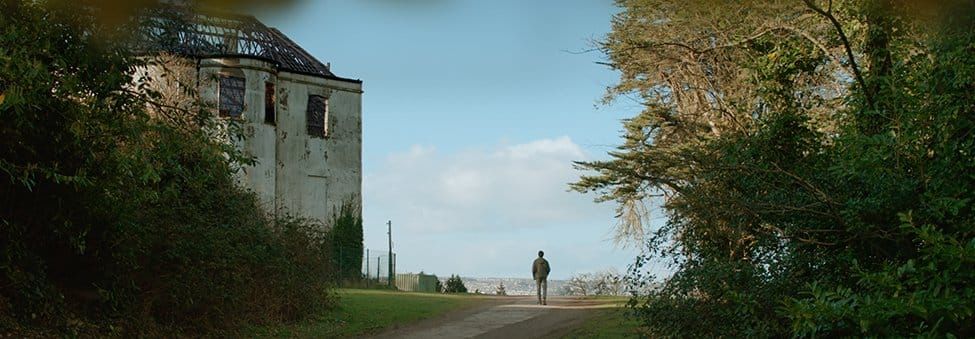 Lone figure walks on a path beside an abandoned building with a damaged roof, flanked by lush green trees under cloudy sky.