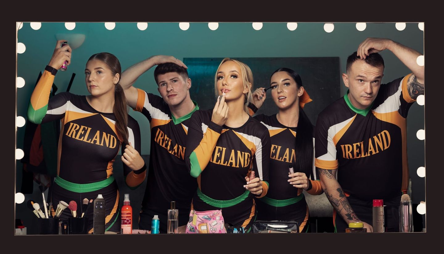 Five Irish cheerleaders in matching black, orange, and green "IRELAND" uniforms getting ready at a lit vanity mirror backstage.