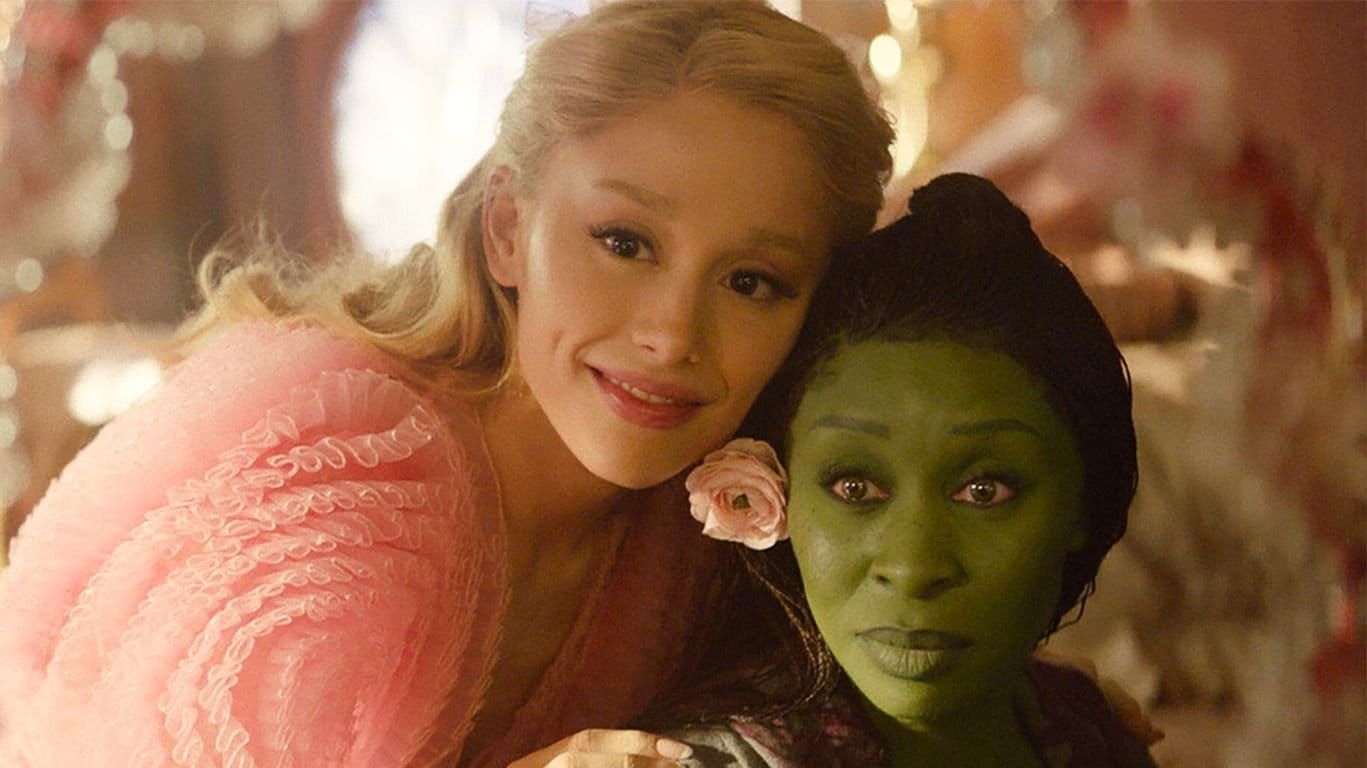 Two women posing cheek-to-cheek; one in a pink ruffled dress with blonde hair, the other with green face makeup and a black hat, from the movie Wicked.