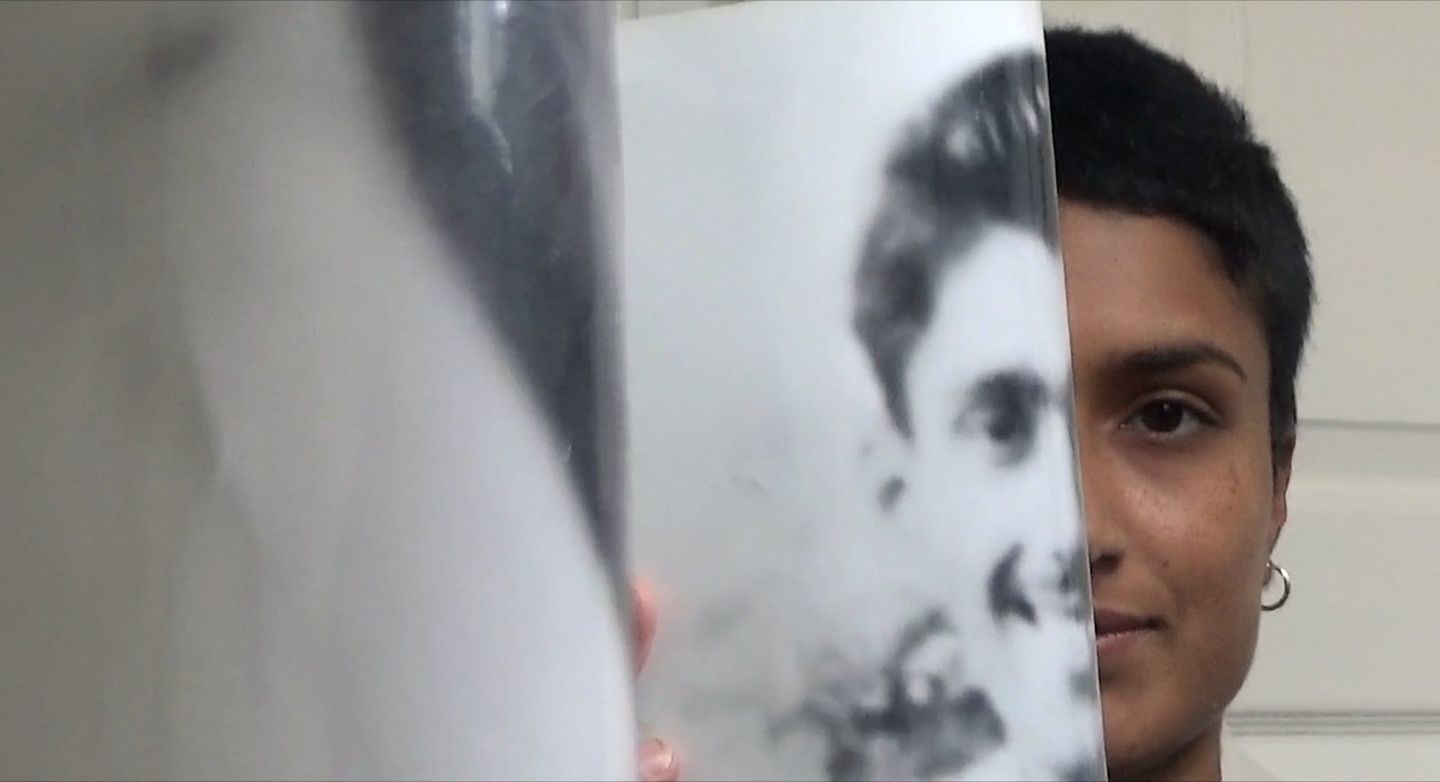 Person with short dark hair and hoop earring peeks from behind a black-and-white printed photograph of someone's face.