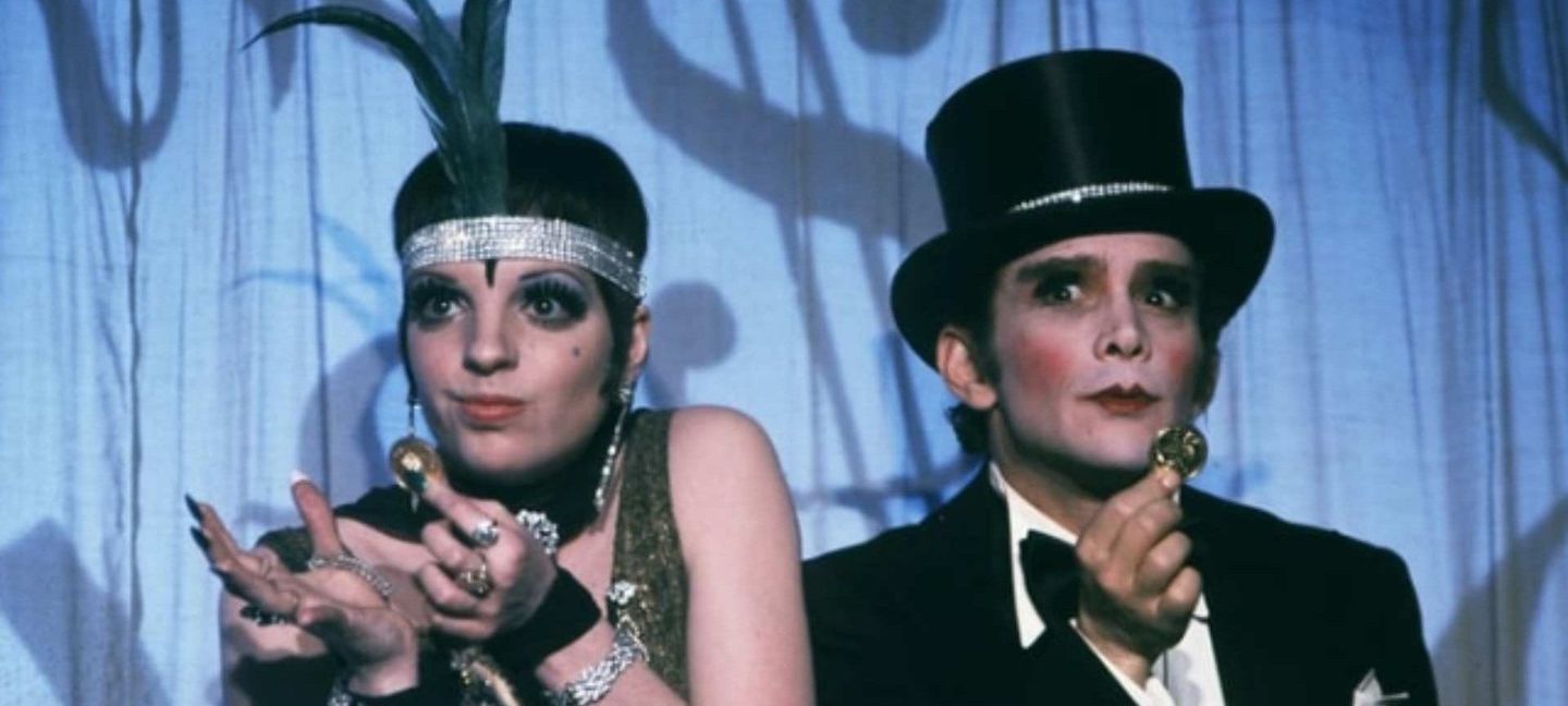 Two cabaret performers in 1920s-style costumes: woman with feathered headband and heavy makeup, man in top hat and tuxedo.