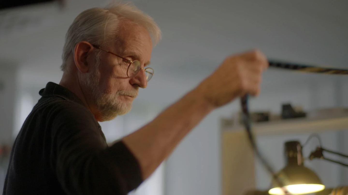 Elderly man with white hair, beard, and glasses reaching up while working at a desk with a brass lamp nearby.