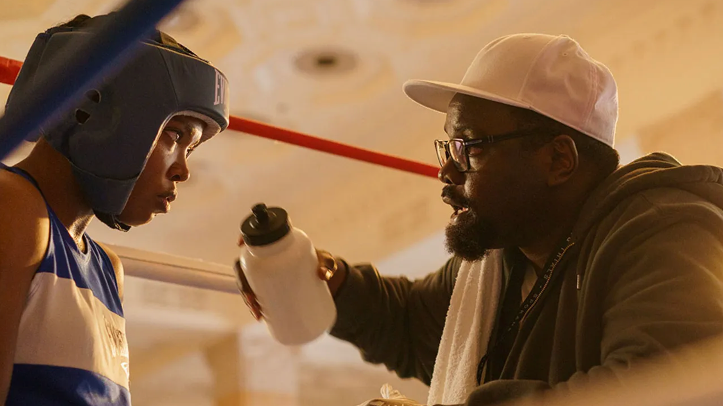 Boxing coach in white cap and glasses offers water bottle to young boxer wearing blue headgear in a ring corner.