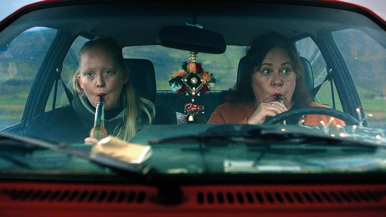Two women sitting in a red car, each sipping from bottles through straws, with a decorative ornament hanging from the rearview mirror.