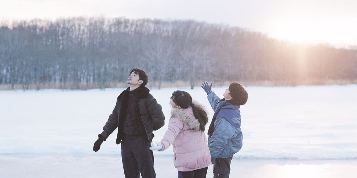 Three people in winter coats standing on a snow-covered field, looking up at the sky, with bare trees and sunset behind them.