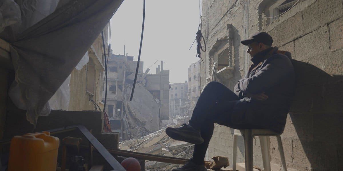 Man sitting on chair overlooking heavily damaged and bombed-out urban buildings, surrounded by rubble and debris.