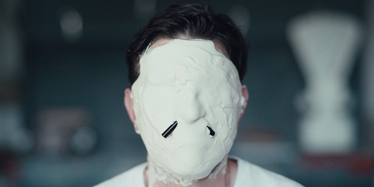 Person wearing a smooth white featureless mask covering their entire face, with two small dark slits near the nose area.