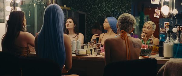 Three women sit at a backstage dressing room vanity with lit mirrors, applying makeup, wearing colorful wigs and costumes.