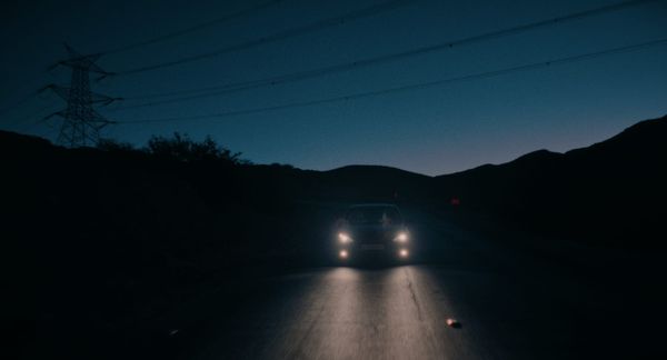 A car travels down a road at twilight with its headlights on, darkness surrounding it.
