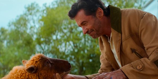 Man in tan jacket smiling down at a small curly-haired brown lamb outdoors with green trees in the background.