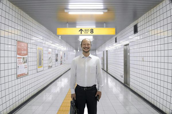 Smiling man in white shirt and dark pants holding a briefcase in a Japanese subway corridor under Exit 8 sign.