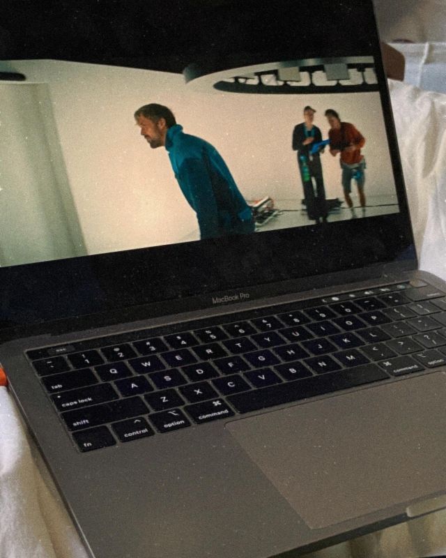 MacBook Pro displaying a movie scene with a man in a blue jacket on a futuristic set, two people visible in background.