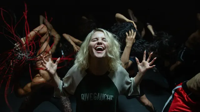 Blonde woman in "Rive Gauche" shirt laughing with outstretched hands, surrounded by dancers in a dark performance space.