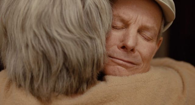 Two elderly people sharing an emotional embrace, one with eyes closed and a gentle smile, the other with gray hair facing away.