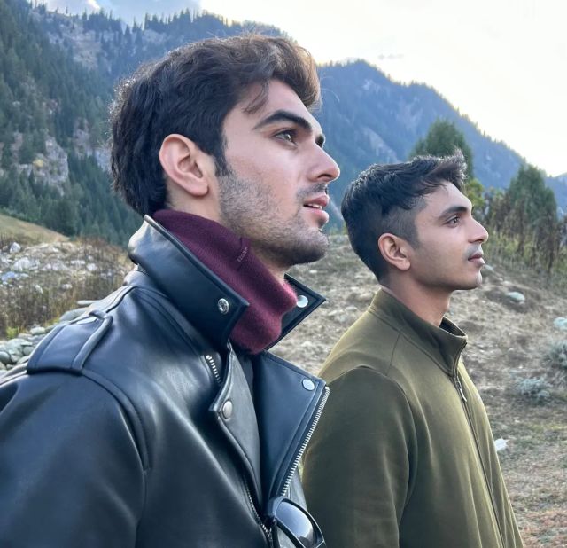 Two young men gazing into the distance on a mountainside, one in a black leather jacket with maroon turtleneck, the other in olive fleece.