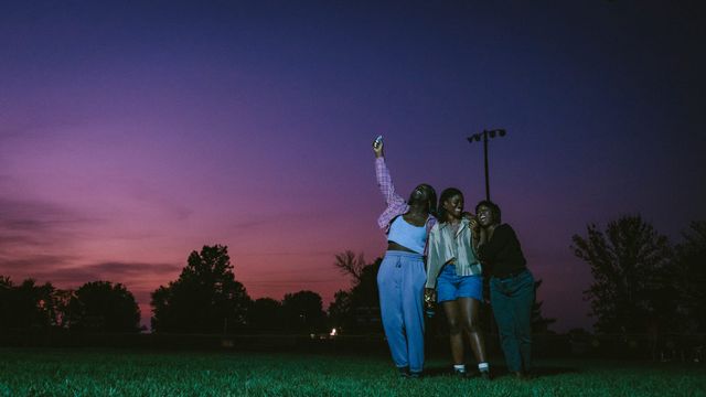 Three friends stand together on grass at dusk, one raising a phone for a selfie, against a vivid purple-pink sunset sky.