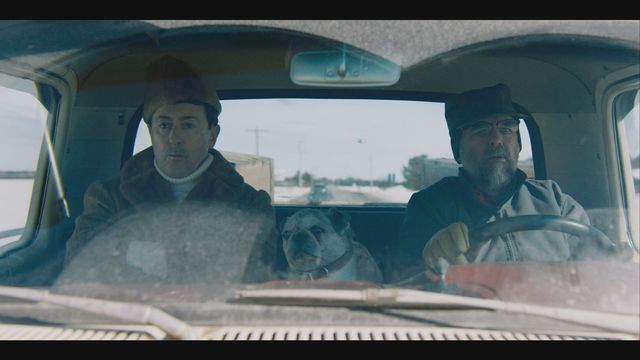 Two men in winter clothing sit in an old truck cab with a bulldog between them, driving on a rural road in cold weather.