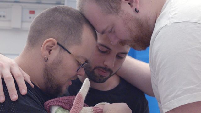 Three men lean close together, tenderly looking down at a newborn baby wrapped in a pink and white knitted blanket.
