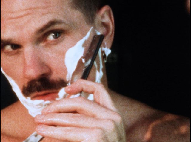 Close-up of a man with a handlebar mustache shaving his cheek with a straight razor, face covered in white shaving cream.