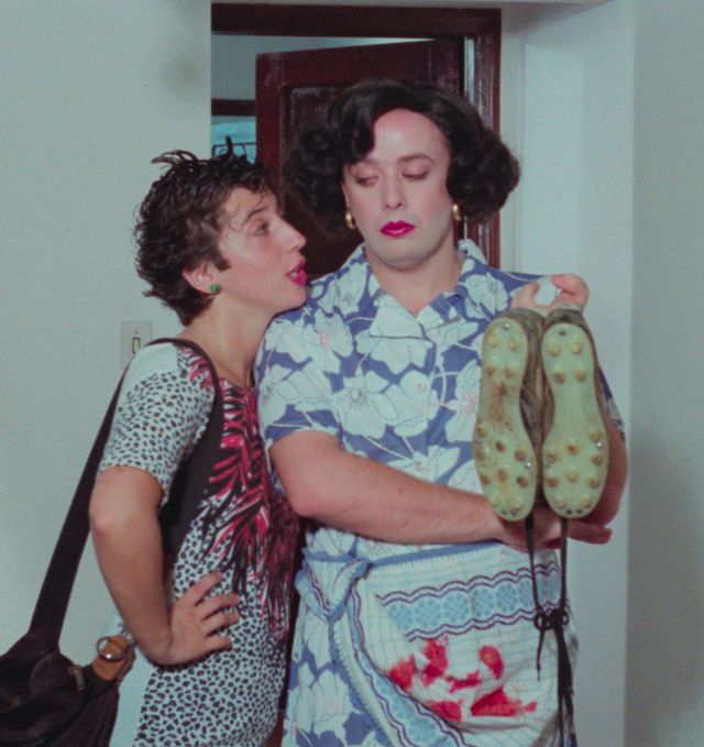 Two people in theatrical costumes and makeup embrace in a hallway, one holding dirty shoes, both wearing patterned tops.