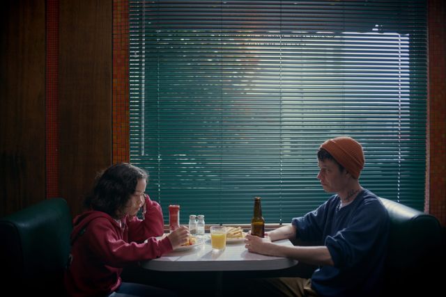 Two people sit across from each other in a diner booth eating fries and drinking, with venetian blinds behind them.