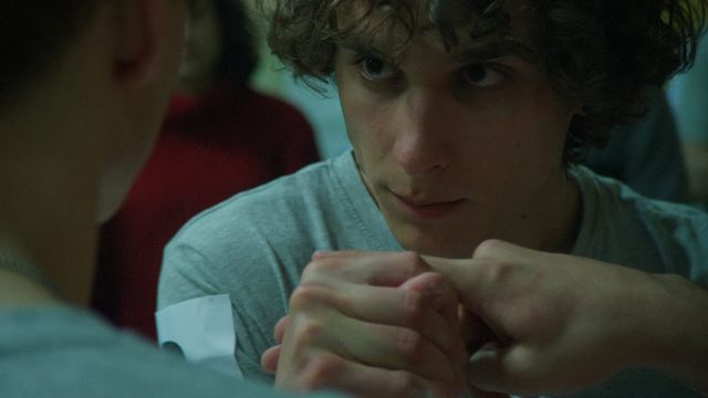 Young man with curly brown hair in a gray t-shirt, leaning forward with clasped hands, looking intently at someone nearby.