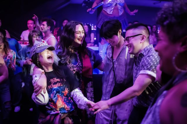 Joyful people dancing at a nightclub, a woman in a tiara and band t-shirt laughing while holding hands with friends.