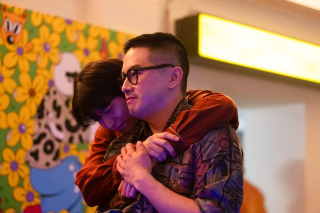 Child hugs a man wearing glasses and a patterned shirt from behind, clasping hands, in front of a colorful cartoon mural.
