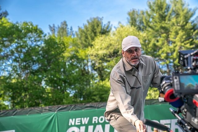Man in white cap and gray jacket leaning forward near a video camera at a New Rochelle Challenger tennis event outdoors.