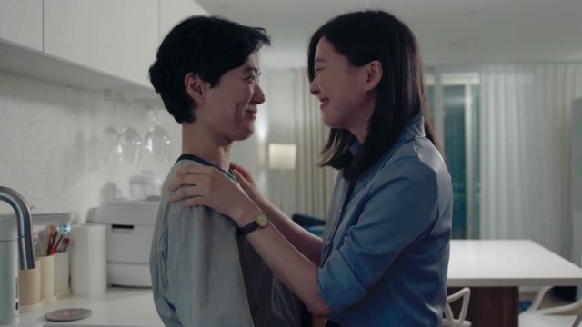 Two people smiling at each other in a modern white kitchen, one with short hair in a gray top, the other in a blue shirt.