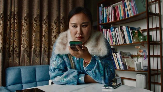 Woman in sparkly blue jacket with white fur collar looks at green smartphone at white desk, bookshelf and curtain behind.