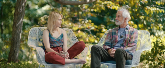 Two people sit in white wicker chairs outdoors: a young blonde woman in a blue top and red pants, and an older gray-haired bearded man in plaid.