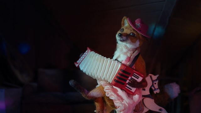 Felt fox puppet wearing a pink cowboy hat and ruffled dress, playing a red accordion against a dark background.