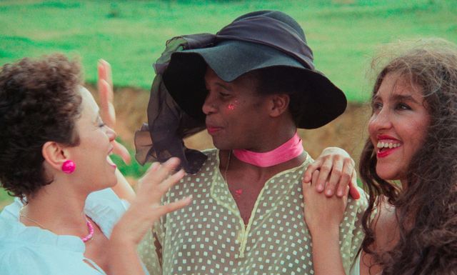 Three friends laughing together outdoors; center person wears a large dark hat, pink choker, and polka-dot shirt with glitter on their cheek.