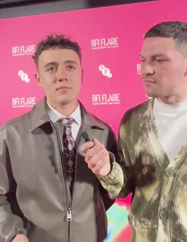 Two young men at BFI Flare London LGBTQIA+ Film Festival, one in leather jacket with tie, other in green cardigan, pink backdrop.