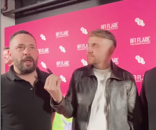 Two men standing in front of a pink BFI Flare film festival backdrop, one speaking into a microphone, the other listening.