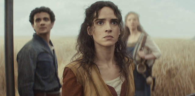 Three people stand in a wheat field; a young woman with curly dark hair looks concerned, flanked by a man and a woman behind her.