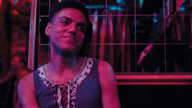 Young man with short curly hair wearing a leopard-print lace-up tank top, smiling in a dimly lit club with pink neon lighting.