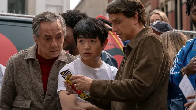 Three men in a crowd: an older Asian man, an Asian teen in a white t-shirt, and a man in a brown jacket showing something.