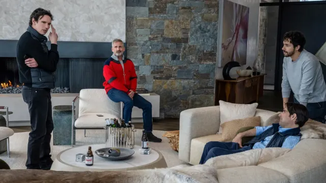 Four men in a modern living room with stone walls and fireplace, drinking beer, one standing, one on chair arm, two on sofa.