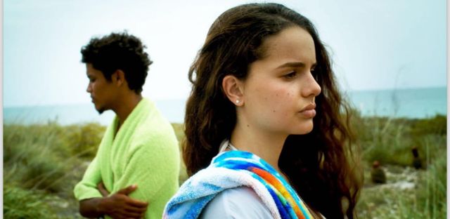 Two teenagers stand on a grassy beach dune looking pensive, wrapped in towels, with the ocean visible behind them.