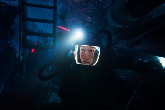 Person in a diving suit with helmet and headlamp in a dark underwater environment near a metal ladder, looking tense.