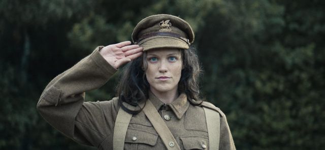 Woman with blue eyes and dark hair saluting in a World War I-era British military uniform and officer's cap outdoors.