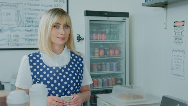 Blonde woman in blue polka-dot apron over white blouse holding cash at a British fish and chip shop counter.