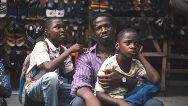 Man sitting with two boys in front of a sandal and shoe display at an outdoor market, looking pensive.